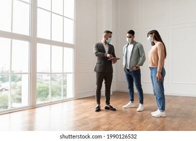 Property Agent In Suit And Protective Medical Mask Showing Blueprints Of New Empty Apartment Flat To Couple During Covid Pandemic Outbreak. Young Man And Woman Visiting And Buying Private House