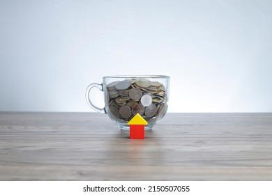 Property investment and financing concept. Selective focus on the house model made from wooden blocks. A glass cup full with coins at the back. Noise is visible due to the texture of the subjects