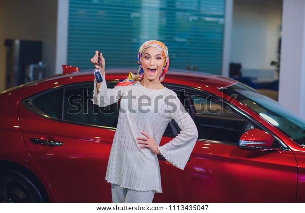 property and people concept - muslim woman\
in hijab with car key over car show background. happy woman taking\
car key from dealer in auto show or\
salon