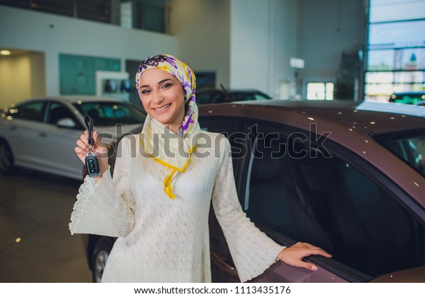 property and people concept - muslim woman\
in hijab with car key over car show background. happy woman taking\
car key from dealer in auto show or\
salon