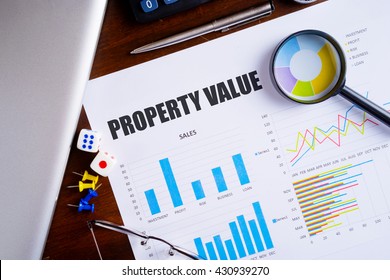 "Property Value" text on paper sheet with magnifying glass on chart, dice, spectacles, pen, laptop and blue and yellow push pin on wooden table - business, banking, finance and investment concept