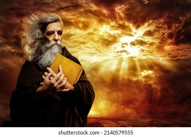 Prophet with Bible. Old Monk with Golden Book praying over Epic Landscape Background. Senior Bearded Man Worship in Black Cloak over Mystery Sunset Sky