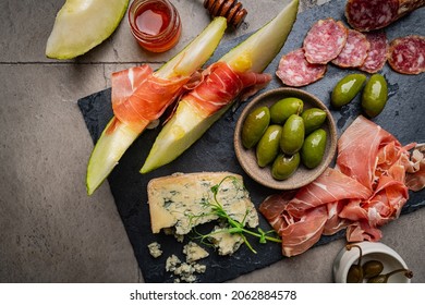 Prosciutto Ham, Slices Of Melon, Brie And Gorgonzola Cheese, Honey And Olives On Gray Background, Top View