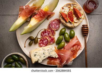 Prosciutto Ham, Slices Of Melon, Brie And Gorgonzola Cheese, Honey And Olives On Gray Background, Top View