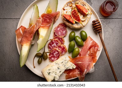 Prosciutto Ham, Slices Of Melon, Brie And Gorgonzola Cheese, Honey And Olives On Gray Background, Top View