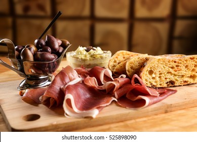 Prosciutto On Wooden Platter With Olives And Backed Bread With Herbs.