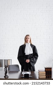 Prosecutor In Black Mantle Looking At Camera Near Desk And White Wall In Office