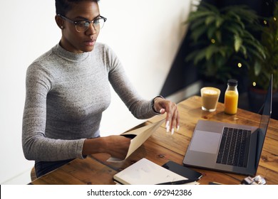 Prosperous Afro American Female Entrepreneur Opening Envelope Of Post Letter Receiving Documentation Via Mail Delivery, Confident Businesswoman In Eyewear Packing Papers With Reports For Sending 