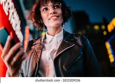 Prosperous party woman with smartphone technology enjoying nightlife in metropolitan city, happy millennial in optical eyewear and modern headphones listening funny music set during evening walk