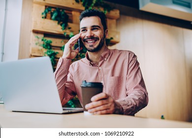 Prosperous Young Male Freelancer Making Telephone Call While Working Remotely On Laptop Computer In Coworking Office , Handsome Smiling Hipster Guy Enjoying Mobile Phone Conversation On Coffee Break