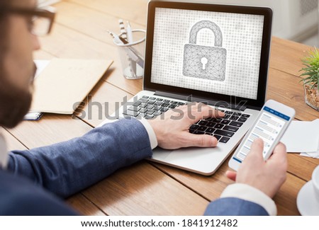 Protect Your Personal Information. Over the shoulder view of man typing on laptop keyboard, holding mobile phone, padlock icon on the computer screen. Data And Cyber Security Concept.