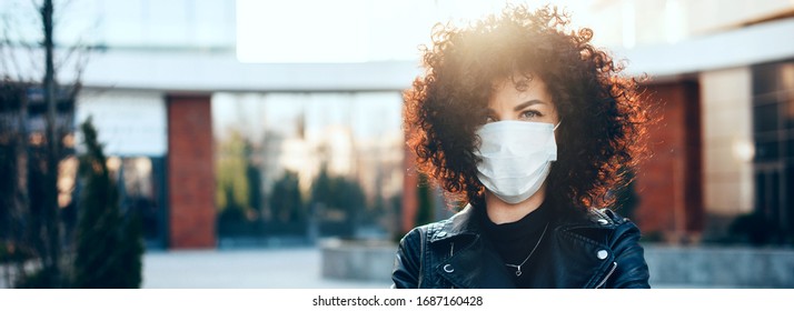 Protected caucasian woman with curly hair is posing in a sunny day while looking at camera and wearing a special white mask - Powered by Shutterstock