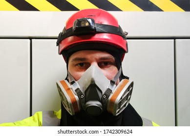 Protection Of Breathing By Employee. Worker In Red Helmet With Respirator On Face Which Prevents From Poor Chemical Smells, Smoke Or Viruses. Gray Metal Wall With Warning Sign Behind Man. Safety Work.