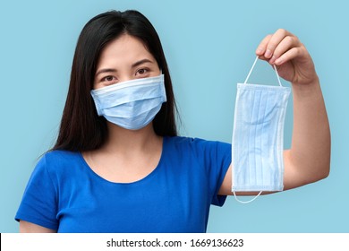 Protection And Epidemic Concept. Young Adult Asian Girl With Covered Nose And Mouth Holding In Hands Safety Face Mask, Standing Isolated On Grey Background