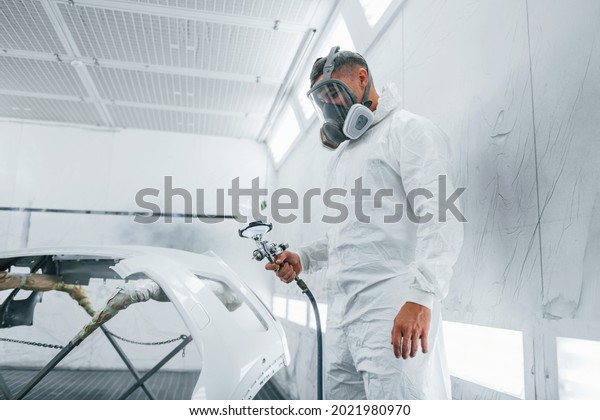 In protective mask. Painting\
the car. Caucasian automobile repairman in uniform works in\
garage.