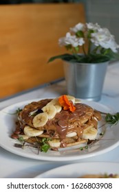 Protein Pancakes With Melted Chocolate Chips And Banana On Them. Pancakes Covered In Edible Flowers In A Cafe Or Restaurant On A White Marble Table.