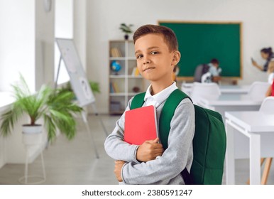 Proud boy first grader with satchel and workbook posing on first day of elementary school getting ready to receive quality assignment standing in classroom with big blackboard. Back to school - Powered by Shutterstock