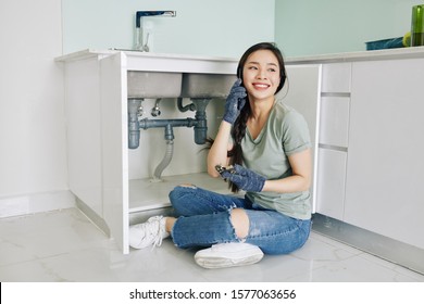 Proud Cheerful Young Asian Woman Sitting On Kitchen Floor And Talking On Phone After Fixing Leaking Sink Pipe