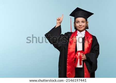 Proud child girl in graduate suit with certificate diploma on light blue background looking at camera, showing promo, pointing finger up, inviting use link. Graduate Celebrating Graduation.