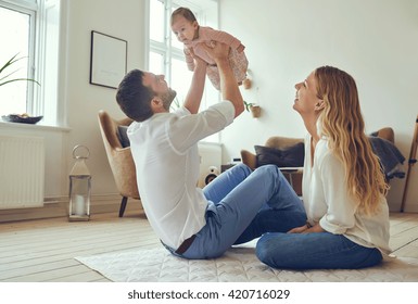 Proud Father Holding His Newborn Baby Daughter In The Air, With Smiling Mother Sitting On The Floor At Home