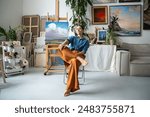 Proud female artist in confident pose sitting on chair, looking at camera. Arrogant woman painter relaxing resting with crossed legs. Brave bold girl among artworks masterpieces in home art studio.