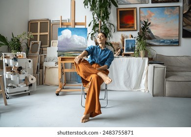 Proud female artist in confident pose sitting on chair, looking at camera. Arrogant woman painter relaxing resting with crossed legs. Brave bold girl among artworks masterpieces in home art studio. - Powered by Shutterstock