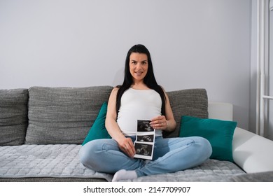 Proud Future Mother Shows Ultrasound Pictures Of Her Unborn Baby. Black-haired Caucasian Pregnant Female Sits With Crossed Legs And Holds Ultrasound Images Between Her Legs.She Leans Against A Pillow.