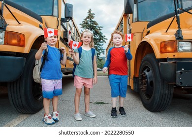 Proud Happy Children Students Boys And Girl Holding Waving Canadian Flags. Education And Back To School In September. Group Of Friends Kids Near Yellow School Bus Outdoors On School Yard. 