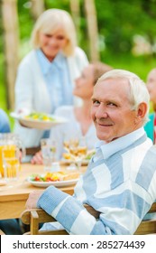 Proud To Have A Big Family. Happy Family Sitting At The Dining Table Outdoors While Senior Man Looking Over Shoulder And Smiling 