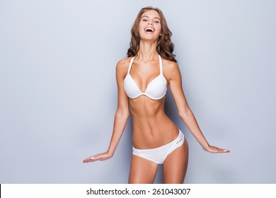 Proud of her perfect body. Attractive young brown hair woman in white lingerie posing against grey background and smiling