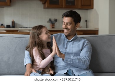 Proud kid girl and happy father giving high five on home couch, hugging, talking, laughing, enjoying leisure. Father giving praise, support to little daughter. Family, parenthood, fatherhood concept - Powered by Shutterstock