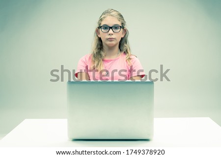 Proud little blond girl seated at her laptop with a deadpan stern expression wearing glasses in a spoof of a scholarly student or businesswoman