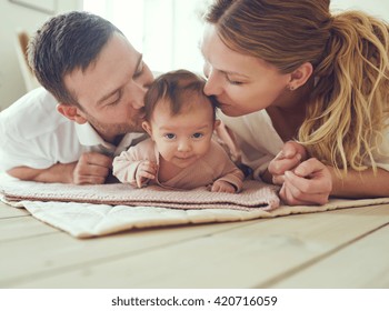 Proud Mother And Father Smiling At Their Newborn Baby Daughter, Lying On The Floor At Home