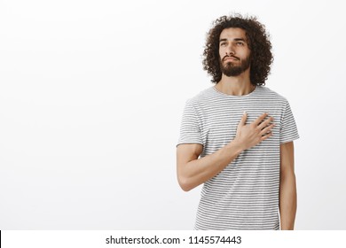 Proud Of My Country. Portrait Of Handsome Thoutful Eastern Man With Beard And Curly Hair, Holding Palm On Heart And Looking Away With Passionate Caring Expression, Being Dedicated To Own Team