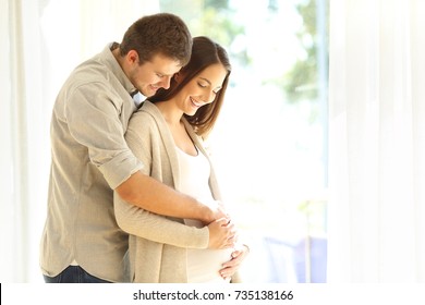 Proud new parents looking at belly of pregnant mother standing beside a window at home