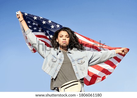 Proud serious young Afro American woman standing on street holding usa flag under blue sky looking at camera. Mixed race lady for democracy, racial equality, fair elections, changes in United States.