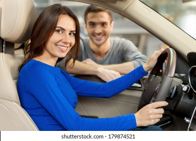 Proud Of Their New Car. Beautiful Young Couple Are Examining A New Car At The Dealership