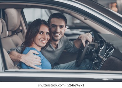 Proud Of Their New Car. Beautiful Young Couple Sitting At The Front Seat Of The Car And Looking At Camera