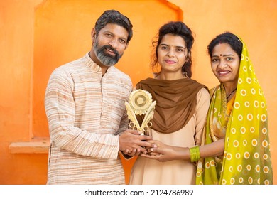 Proud Traditional Indian Parents With Young Daughter Holding Winning Prize Celebrating Victory. Happy Young School Girl Showing Trophy Standing With Her Father And Mother.