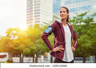 Proud Woman Listening Music With Earphones While Standing And Resting After Running Outdoors. Mature Lady Wearing Jacket And Standing After Morning Run In Urban City. Satisfied Runner Looking Away.