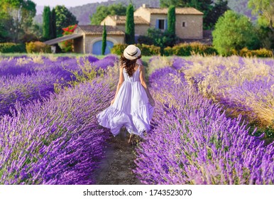 Provence, France. Charming Young Woman In Blooming Lavender Fields At Background Of Beautiful Traditional French Provencal House.  Back View Of Lovely Lady Wearing Waving Boho Chic Purple Dress, Hat.