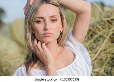 Provincial Young Woman Of Blonde, Of Eastern European Descent In A White Dress Posing Against A Background Of Haystacks. A Young Girl From The Village, A Haystack, A Farmer Girl, National Economy.