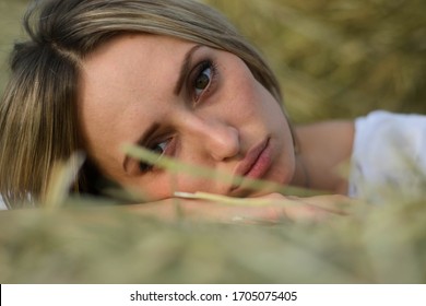 Provincial Young Woman Of Blonde, Of Eastern European Descent In A White Dress Posing Against A Background Of Haystacks. A Young Girl From The Village, A Haystack, A Farmer Girl, National Economy.