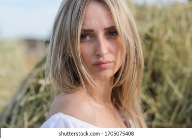 Provincial Young Woman Of Blonde, Of Eastern European Descent In A White Dress Posing Against A Background Of Haystacks. A Young Girl From The Village, A Haystack, A Farmer Girl, National Economy.