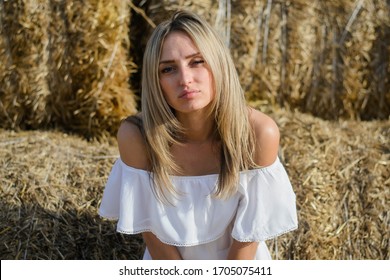 Provincial Young Woman Of Blonde, Of Eastern European Descent In A White Dress Posing Against A Background Of Haystacks. A Young Girl From The Village, A Haystack, A Farmer Girl, National Economy.