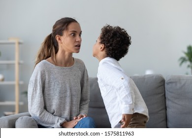 Provocative mixed race little kid demonstrating impolite disrespectful behavior to angry mother, sitting on sofa in living room at home. Irritated mom talking strictly to naughty son, family conflict.