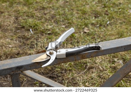 Pruning shear tool resting on a metal fence with grass in background, for gardening, plant care, and agricultural work concept.
