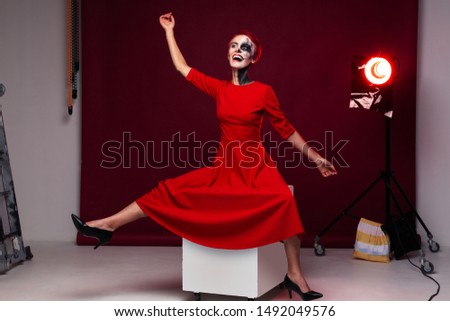 Psycho girl sitting on a cube in strange pose in red dress and with art makeup. Backstage in studio concept.