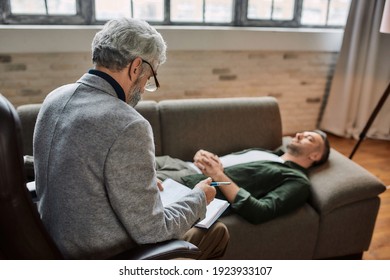Psychoanalyst making notes in notepad during appointment with young caucasian man resting on sofa with closed eyes. Psychotherapy concept