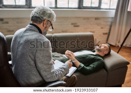 Psychoanalyst making notes in notepad during appointment with young caucasian man resting on sofa with closed eyes. Psychotherapy concept